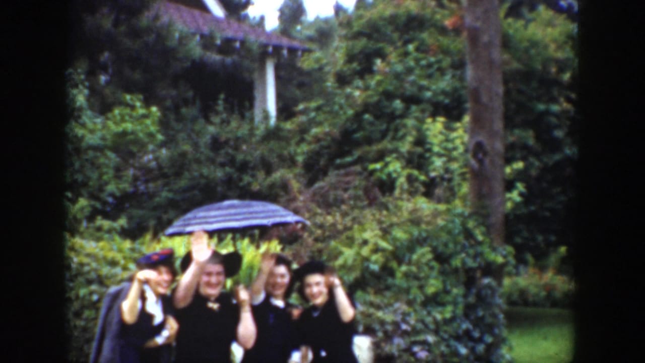Adults in a 1939 Wyoming garden scene smile and wave toward the camera, grouped beneath a striped umbrella among dense greenery.