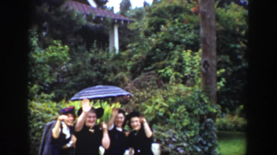 Adults in a 1939 Wyoming garden scene smile and wave toward the camera, grouped beneath a striped umbrella among dense greenery.