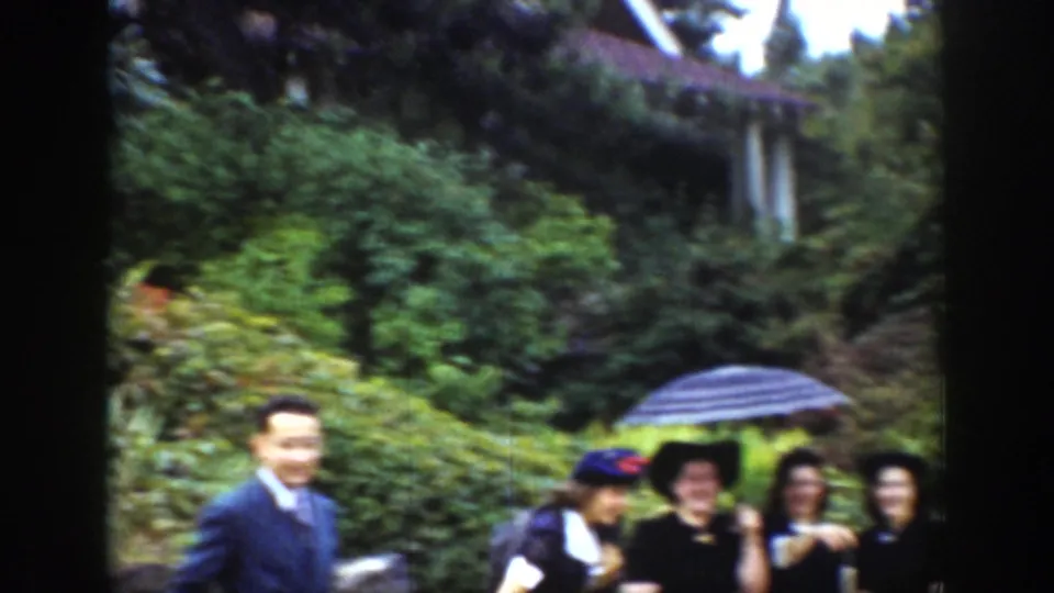 A man in a suit appears beside women under a striped umbrella in the Wyoming garden, with greenery and a house roofline behind them.