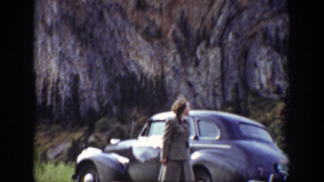 A woman in Wyoming in 1939 appears beside a rounded 1930s sedan parked below steep rocky cliffs.