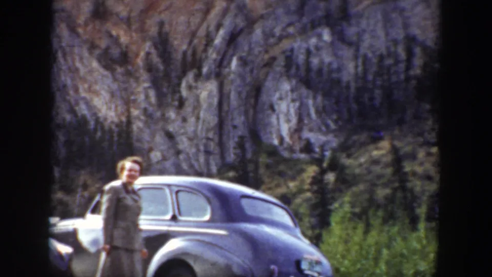 The traveler stands near a dark 1930s sedan with Wyoming cliffs and evergreens behind her.