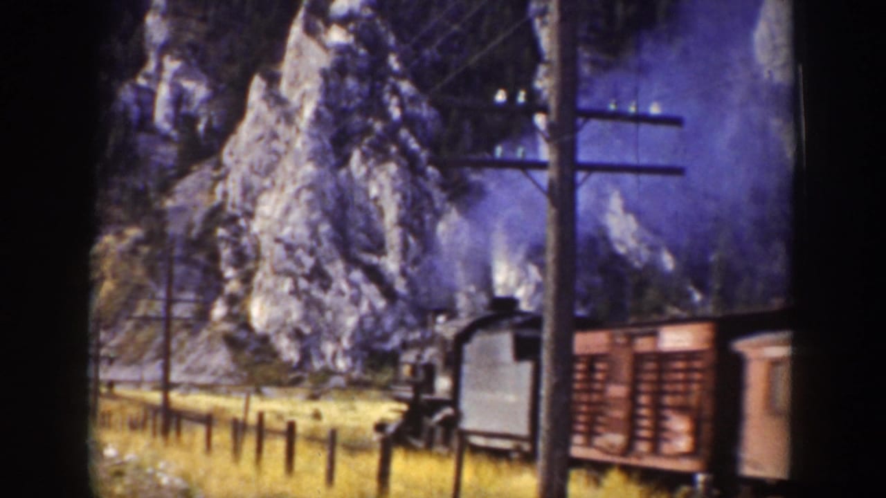 Steam locomotive and cars passing below rocky mountains and utility poles.