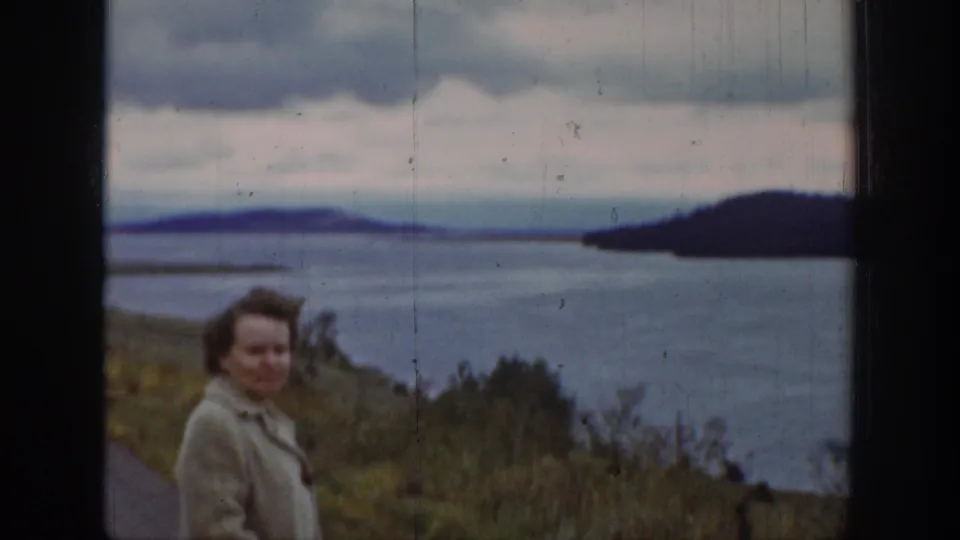 A woman in a light coat faces the camera beside a 1939 Wyoming lake view, with roadside vegetation, hills, and cloudy sky behind her.