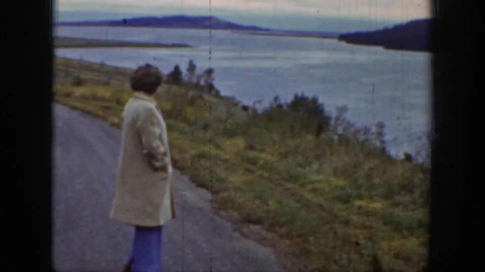 In Wyoming 8mm footage from 1939, a woman in a light coat stands turned away on a rural road above a broad river and low hills.