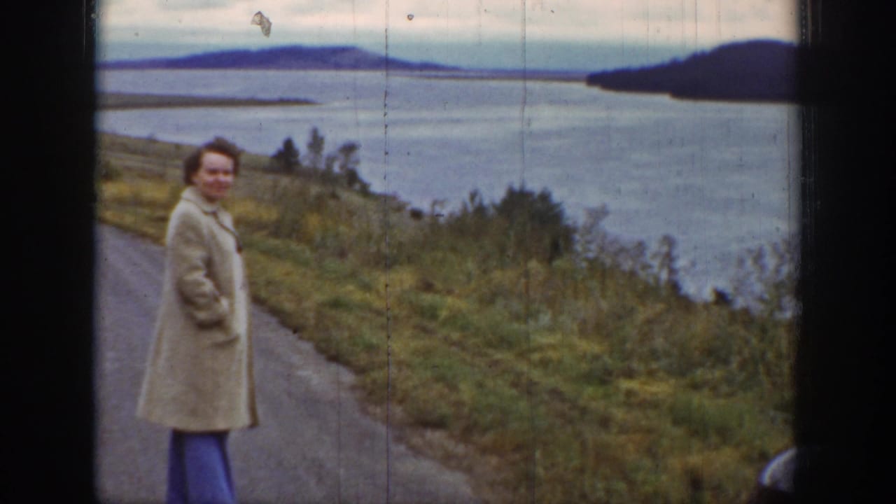 Woman in a light coat smiles toward the camera on a road beside water and hills.
