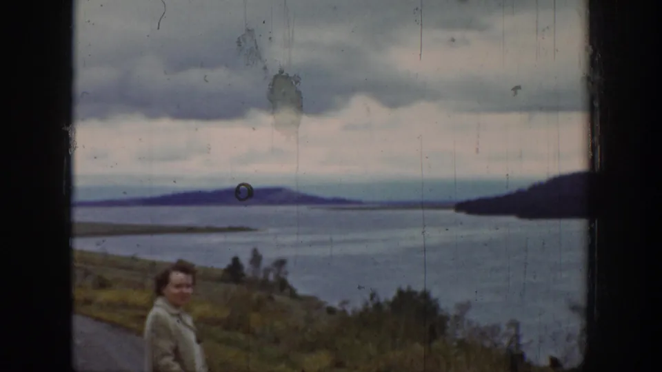 A 1939 Wyoming home-movie frame places the woman low in the composition, with a broad river, dark shoreline, low hills, and cloudy sky above.