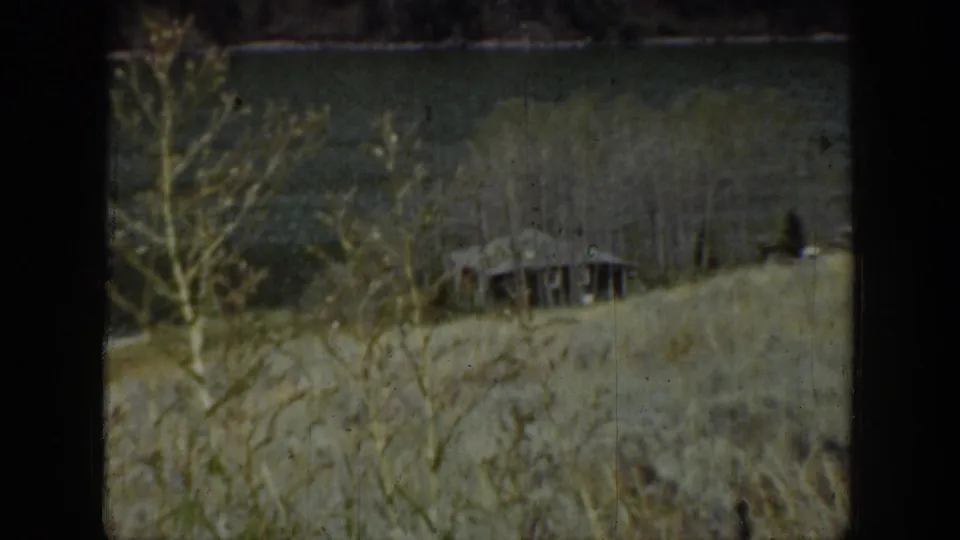 1939 Wyoming home-movie frame showing a grassy lakeside slope, bare trees, and a small rustic wooden hut near the water.