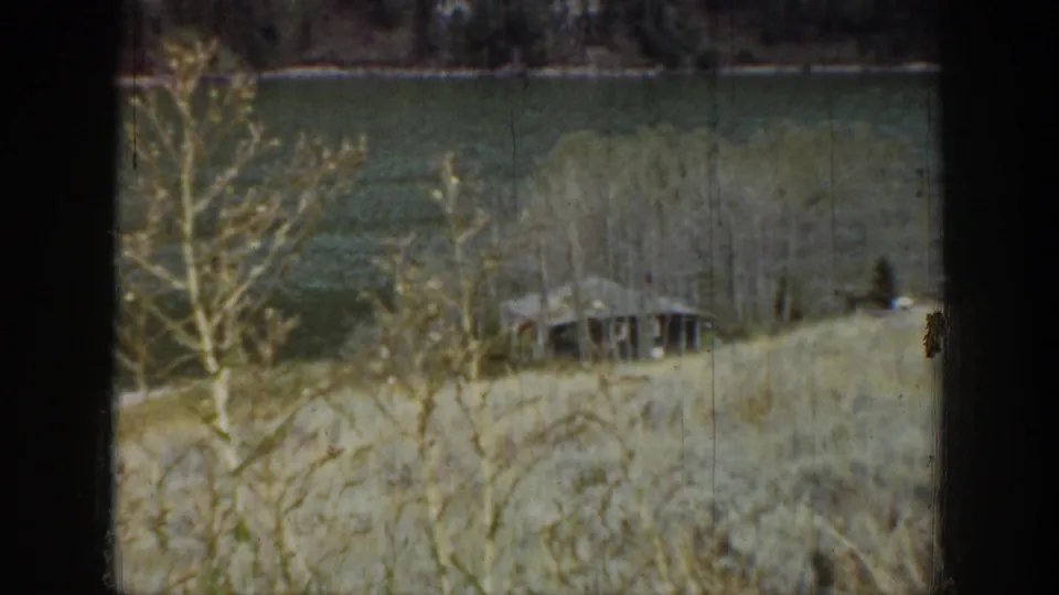 Soft 1939 Wyoming 8mm frame with bare branches in front of a small lakeside hut and muted water beyond.