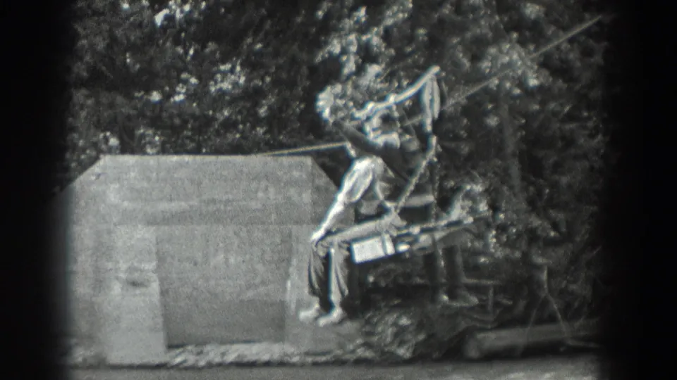 A 1940 Washington home-movie frame shows a man on a suspended cable seat with another figure nearby in the wooded background.