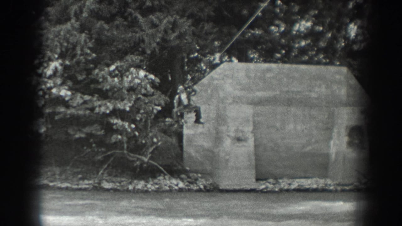 Two people hang from a rope beside a concrete structure near a river and trees.