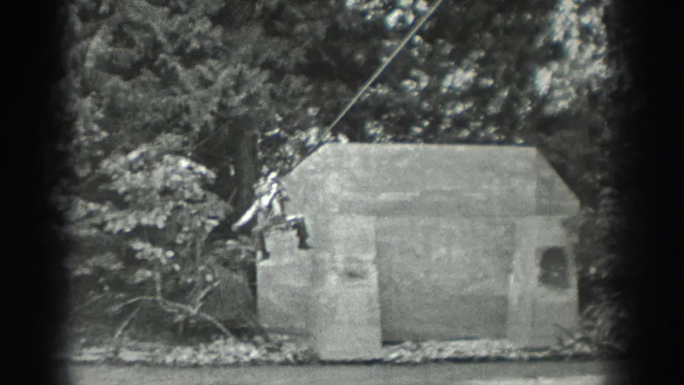 1940 Washington home-movie frame with two distant figures on a rope next to a concrete structure by a wooded riverbank.