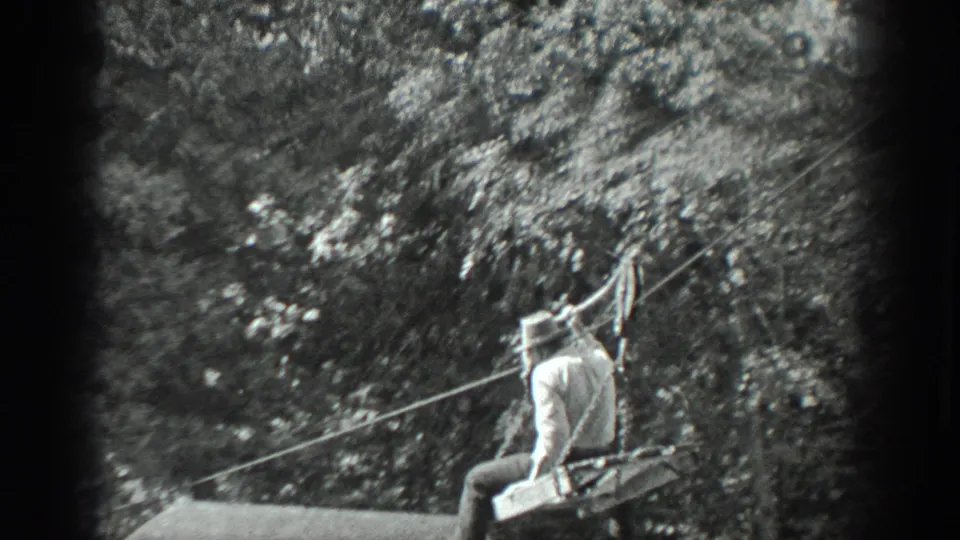 A rear view shows the man seated on the cable apparatus as it travels across the water toward dense trees and brush.