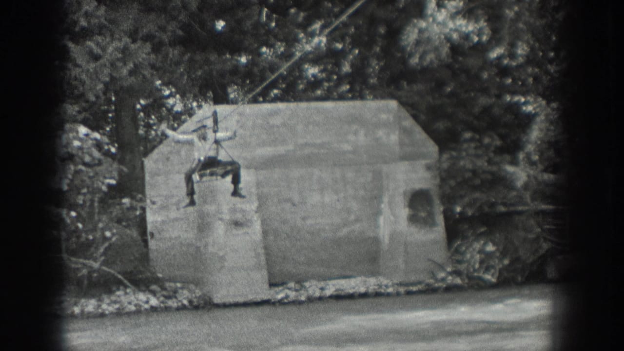 Man sits or hangs on a cable above a concrete structure by a stream.