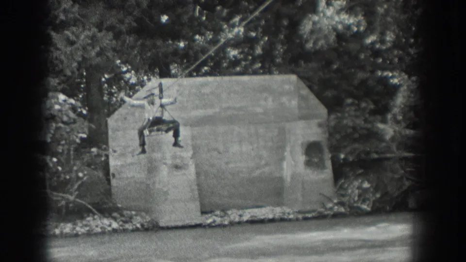 1940 Washington home-movie frame with a man suspended on a cable above a concrete structure next to moving water and trees.