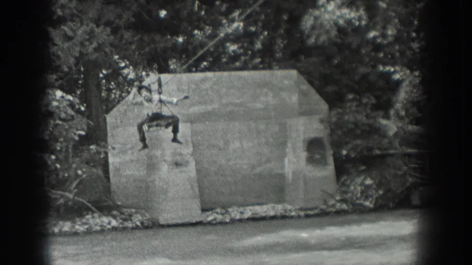 The man stays on the cable over the concrete structure and stream, framed by trees in black-and-white 8mm footage.