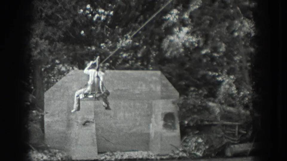 Black-and-white 8mm frame from Washington showing the rider passing over concrete supports in a wooded river area.
