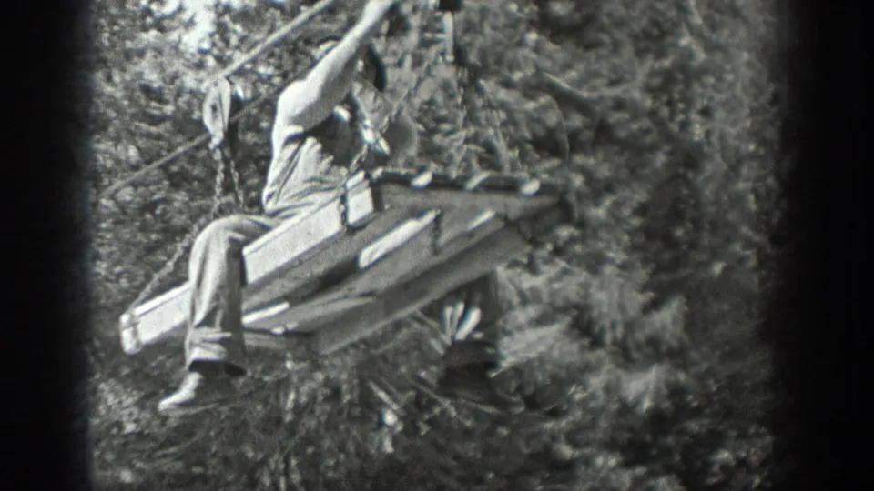 Black-and-white 8mm frame from Washington, 1940, showing a man seated on stacked logs or boards beside a cable in the woods.
