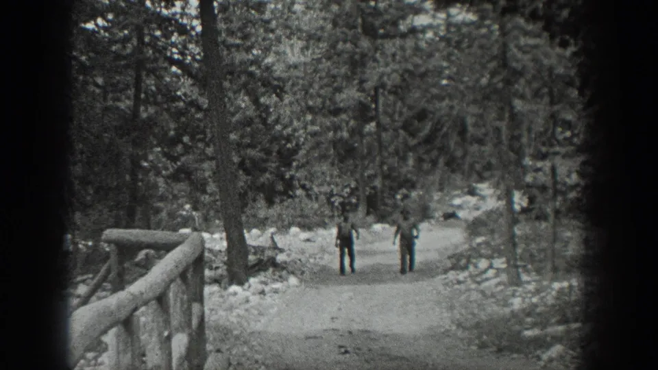 Black-and-white 8mm frame from Washington, 1940, showing two men on a wooded trail near a rustic railing and tall trees.