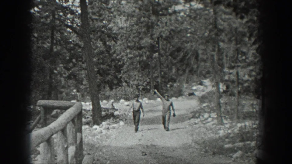 The two men continue toward the camera on a wide wooded path, with a log railing and dense trees visible around them.