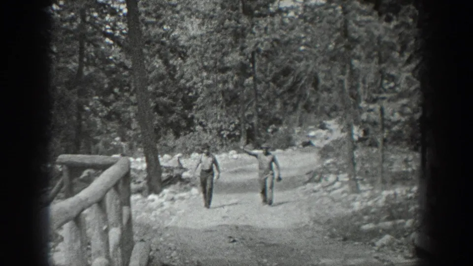 In 1940 Washington home-movie footage, two adult men walk toward the camera on a tree-lined path near a rustic wooden bridge.