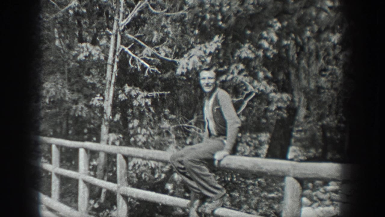 An adult man in period clothing sits on a rustic bridge rail and faces the camera in a wooded Washington setting.