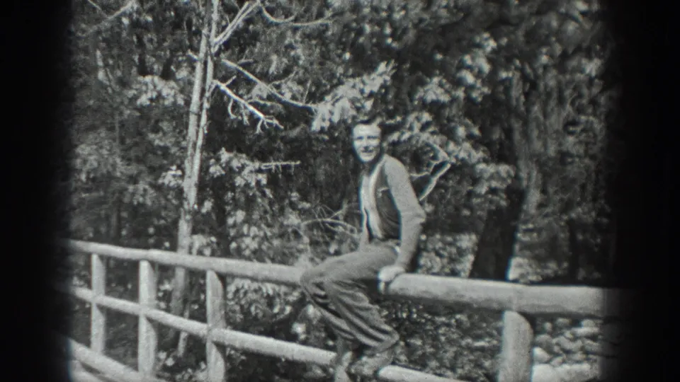 An adult man in period clothing sits on a rustic bridge rail and faces the camera in a wooded Washington setting.