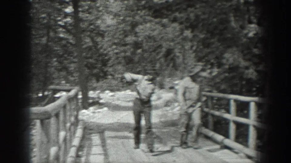 Two adult men appear on or beside a rustic bridge, one closer to the camera, in grainy black-and-white outdoor footage.
