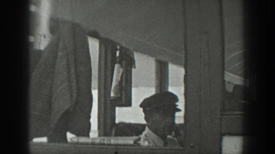 A man in a flat cap sits by the boat windows, with hanging fabric and cabin framing visible in the foreground.