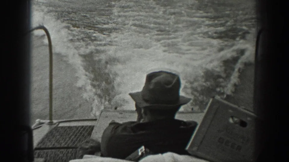 The man remains seated on the moving boat in Washington, with a marked box nearby and turbulent river water behind the stern.