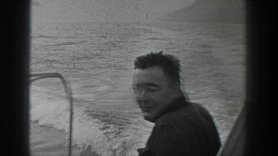 An adult man aboard a moving boat smiles toward the camera, with open water and soft shoreline shapes in the background.