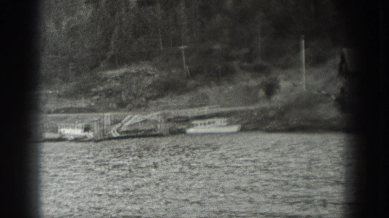 1940 Washington waterfront view with a wooden dock, moored small boats, rocky bank, trees, and utility poles.