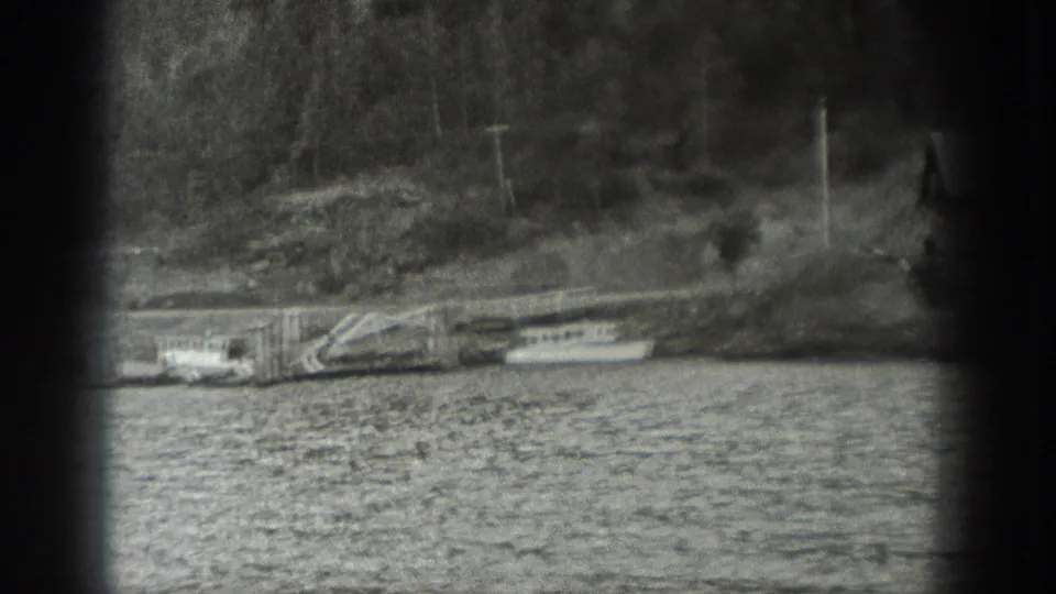 1940 Washington waterfront view with a wooden dock, moored small boats, rocky bank, trees, and utility poles.