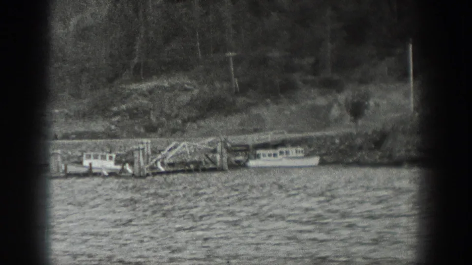 Black-and-white 8mm frame from Washington showing a pier, small boats, water, brush, and a forested shoreline.