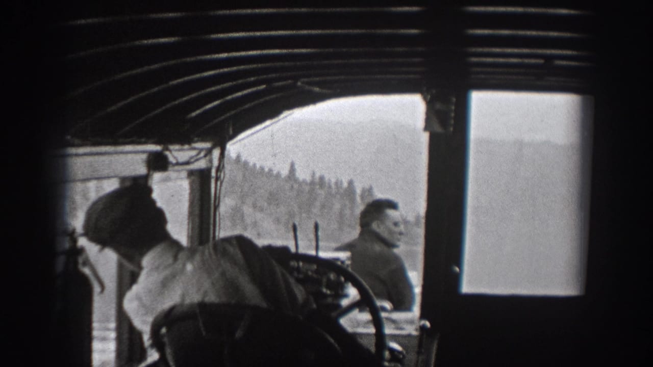 Men sit or stand inside a boat cabin with water and forested shoreline visible through the windows.