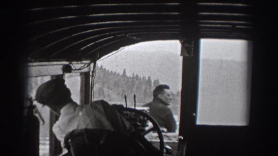 Inside a boat cabin in Washington, men look outward as water and forested shoreline pass beyond the windows.
