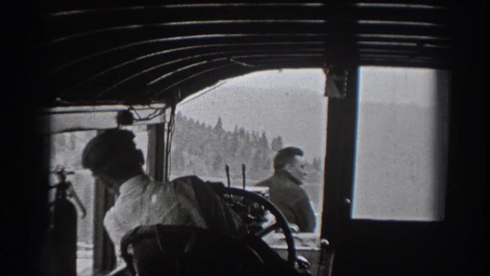 Black-and-white 8mm view from inside the boat cabin, with men near the controls and shoreline outside.