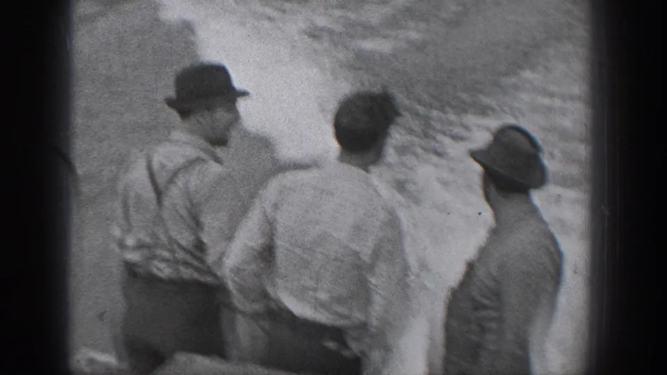 1940 Washington home-movie frame showing three men in hats and shirts gathered near a rough shoreline surface.