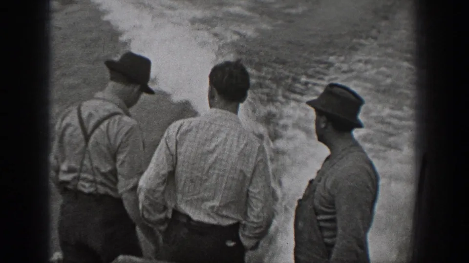 1940 Washington home-movie frame showing adult men in hats, shirts, and suspenders standing on a boat beside churning water.