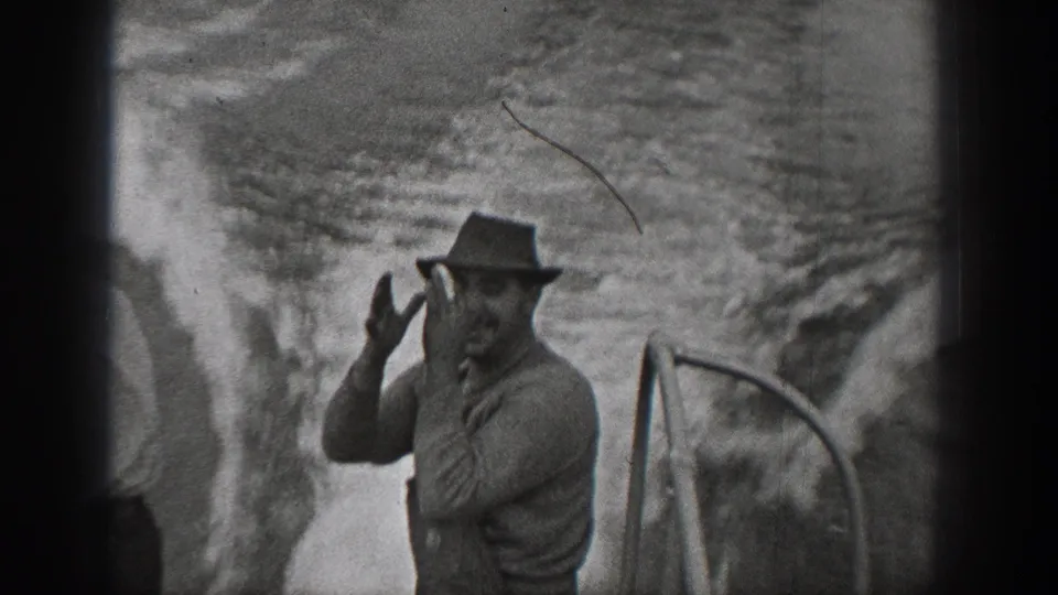 1940 Washington 8mm frame of an adult man in a brimmed hat raising his hands near the boat rail as water churns behind him.