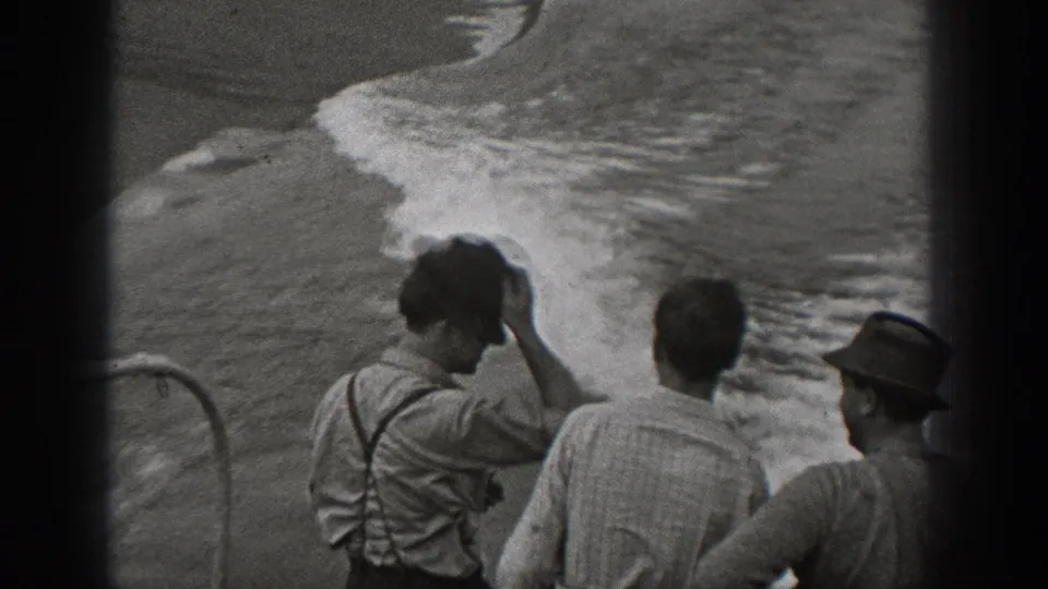 Several adult men, including one in suspenders, watch rough water from a ship deck in black-and-white 8mm footage.