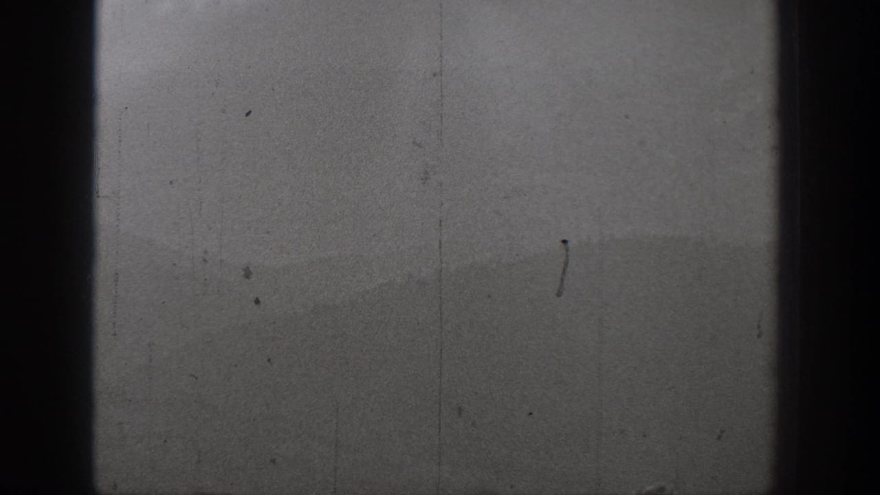 Soft black-and-white 8mm view of water and a hazy mountain or shoreline form, with no close figures visible in this frame.