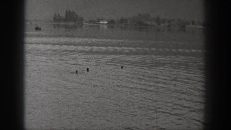 1940 Washington home-movie frame showing a distant swimmer in a lake, with rippled water, shoreline trees, and dark vertical posts in front.
