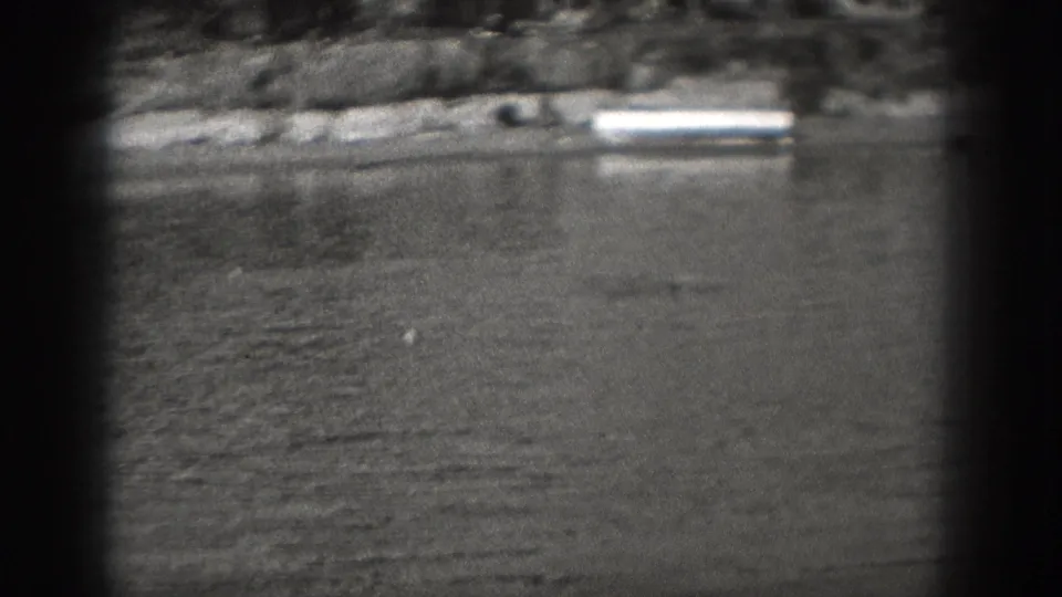 Black-and-white 8mm frame showing open lake water, a distant shore with low structures, and a small swimmer or surface detail near the waterline.