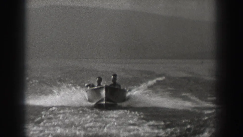 Two men continue across the Washington river in a small motorboat, seen in grainy 8mm footage with a broad wake behind them.