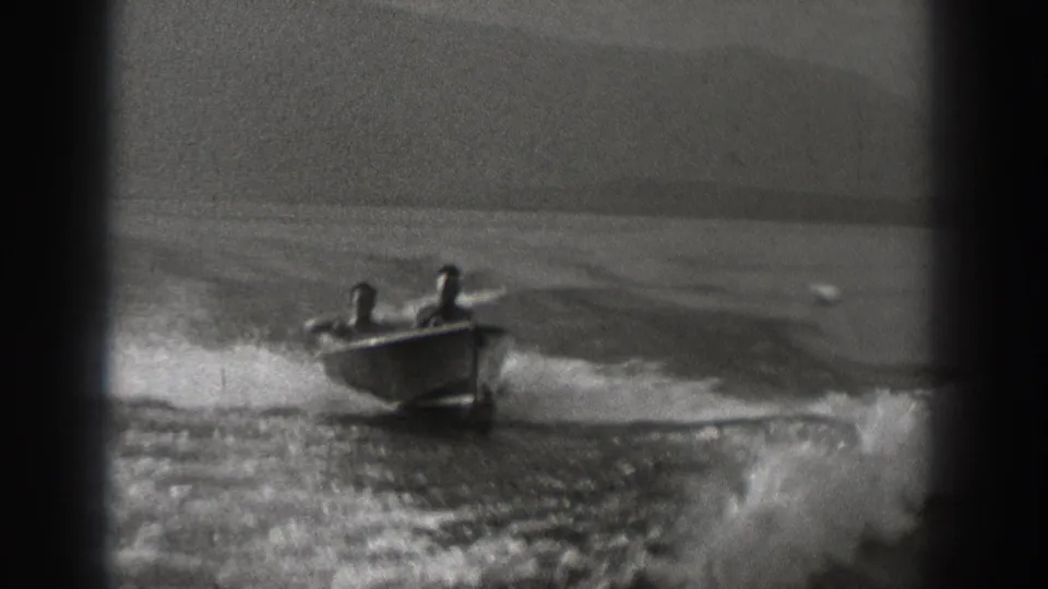The small motorboat moves toward the camera across a broad body of water, with two riders visible in soft 8mm focus.