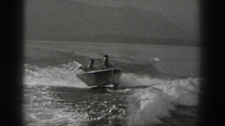 A small open boat carrying two young men crosses the frame, throwing spray across the water surface.