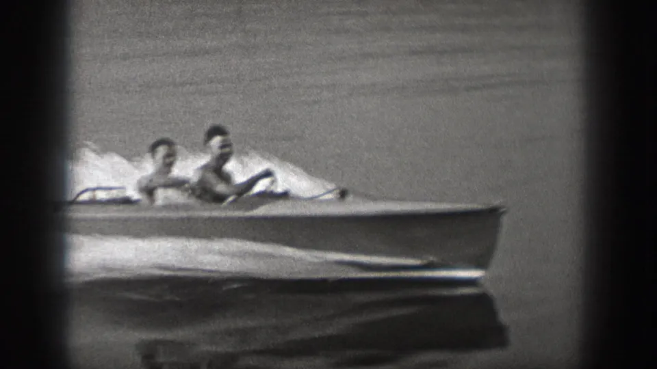 Side view of two young men in a small motorboat, seen in black-and-white 8mm footage on open water.