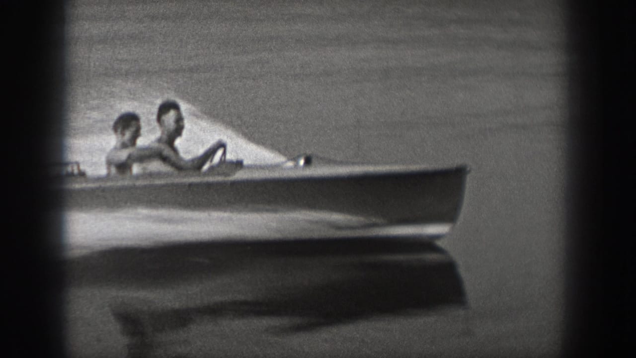 Black-and-white 1940 Washington 8mm frame showing two riders in a small motorboat moving across calm lake water.