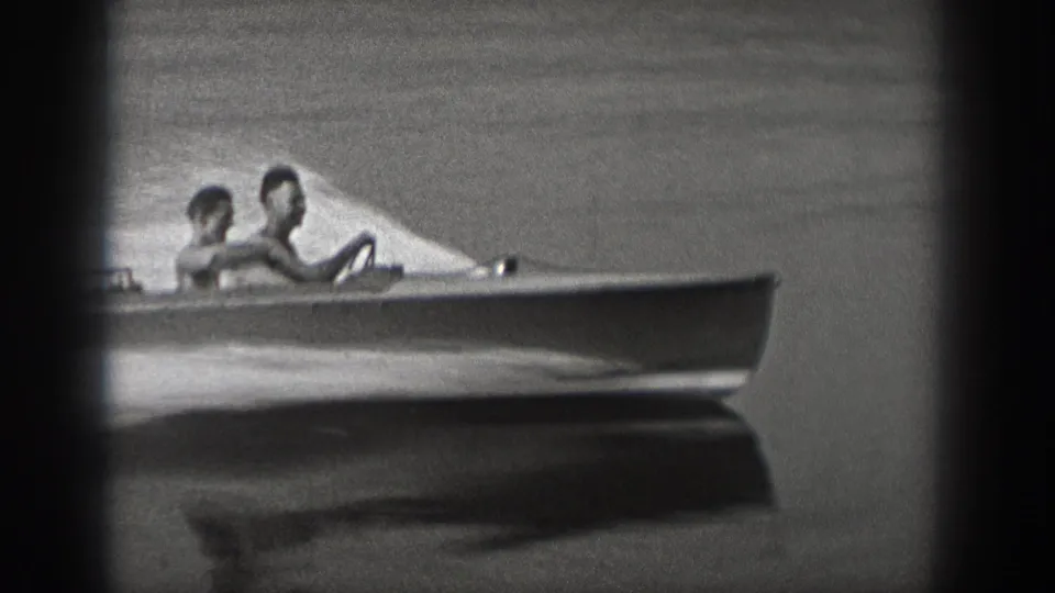 Black-and-white 1940 Washington 8mm frame showing two riders in a small motorboat moving across calm lake water.