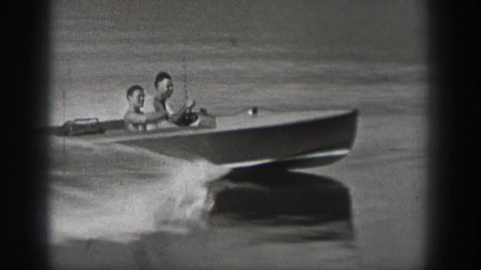 Black-and-white 8mm frame from Washington showing two riders in a speedboat, with water spray and a distant lake surface behind them.